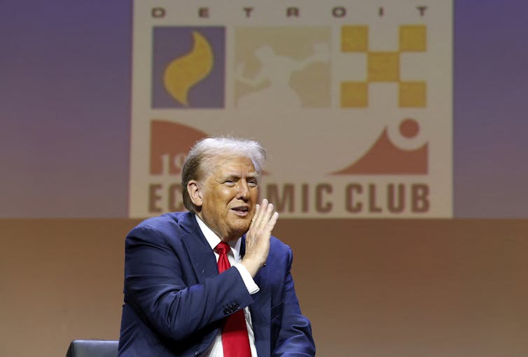 A man in a blue suit and red tie shields his mouth as he speaks