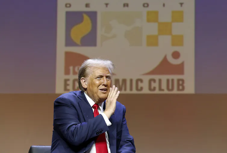 A man in a blue suit and red tie shields his mouth as he speaks