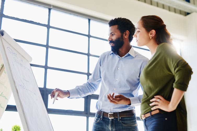 A racialized man and a white woman look at a white board in a brightly lit meeting space