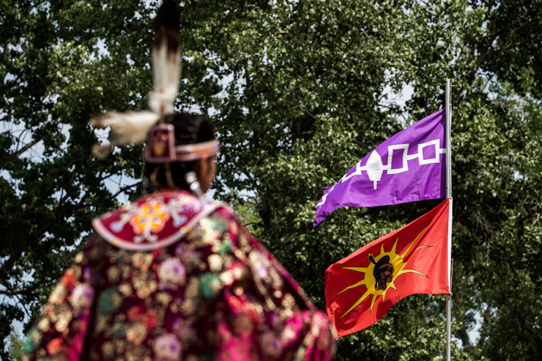 A person in Indigenous American dress stands in front of a flagpole with two flags flying.