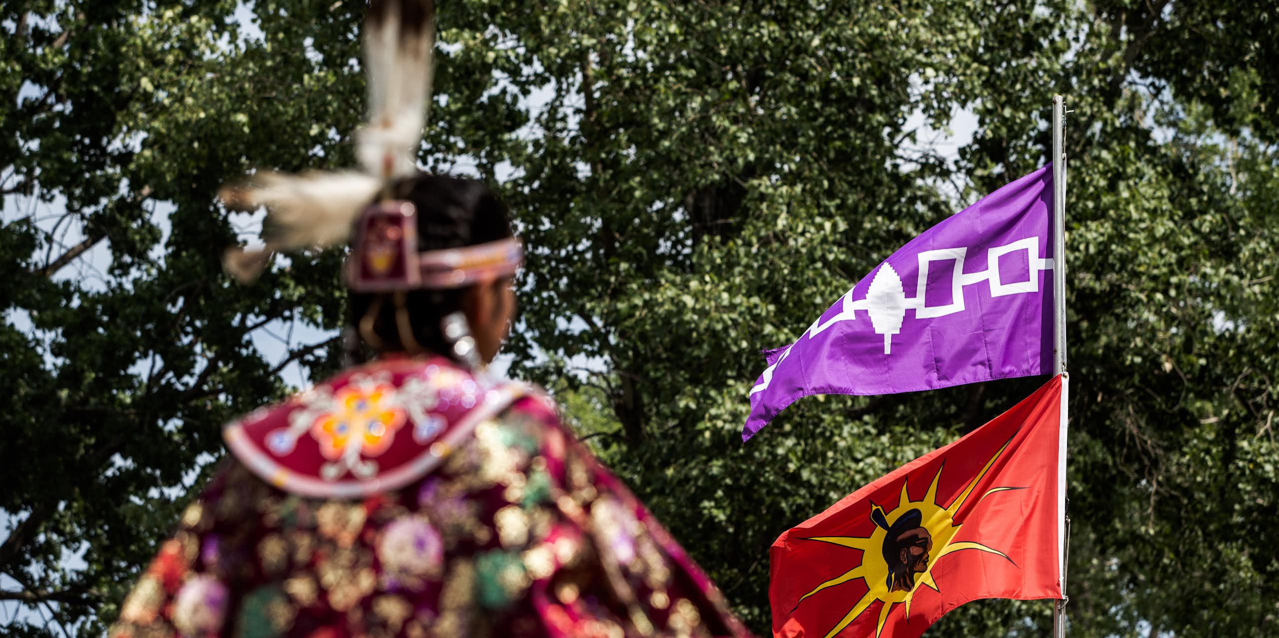 A person in Indigenous American dress stands in front of a flagpole with two flags flying.