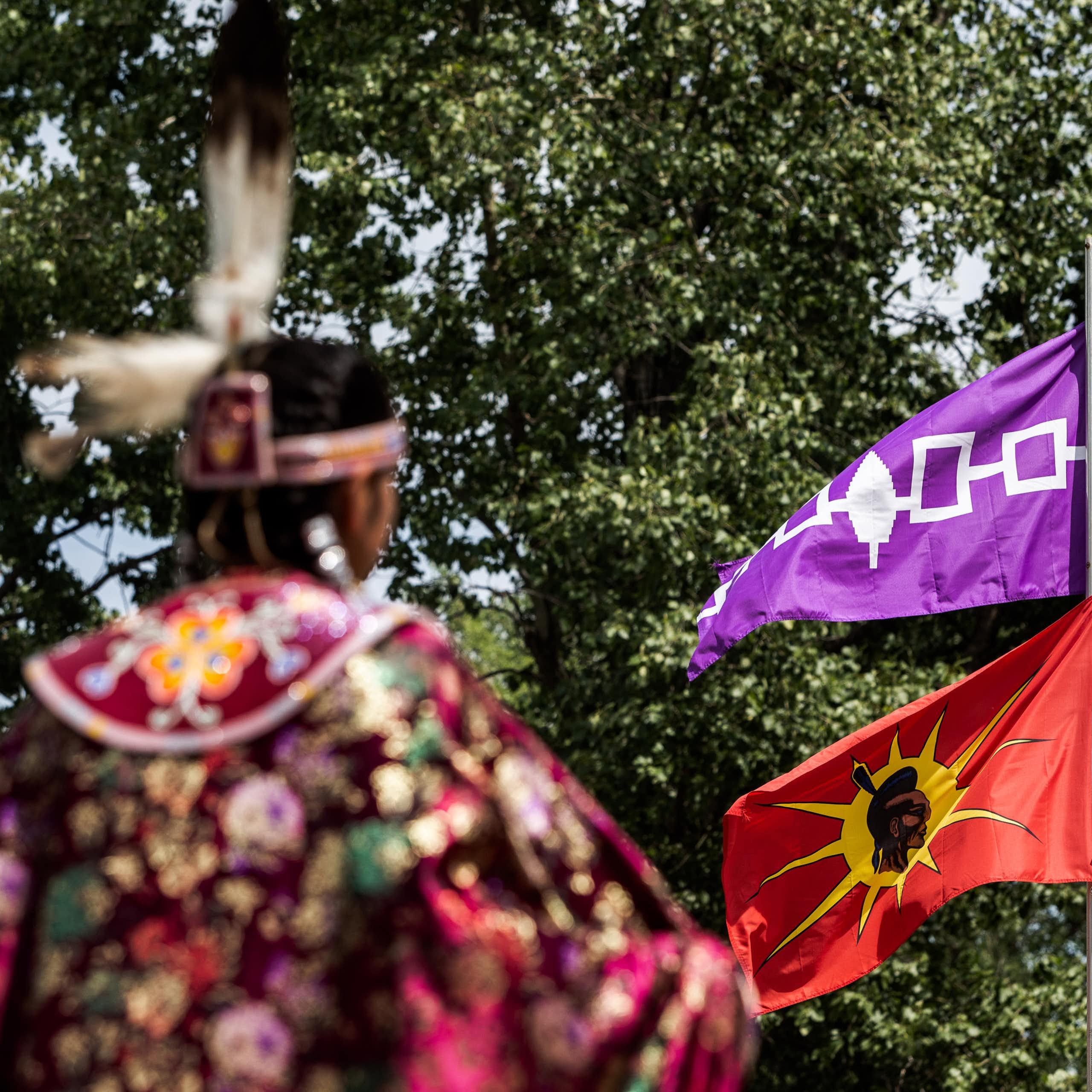 A person in Indigenous American dress stands in front of a flagpole with two flags flying.