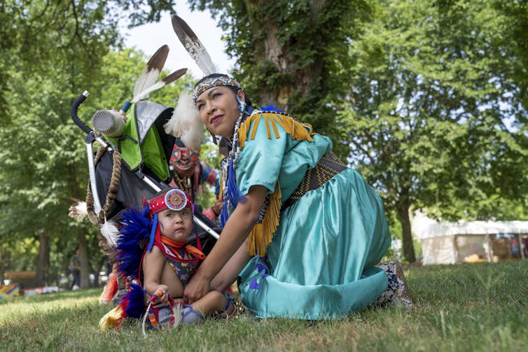A woman wearing a dress with a beaded belt and a beaded headband holding a feather dresses a young child in similar clothing.