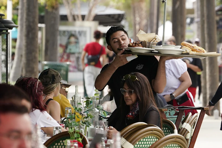 A server balances a large load of food on a tray above his customers' heads in an outdoor dining area.