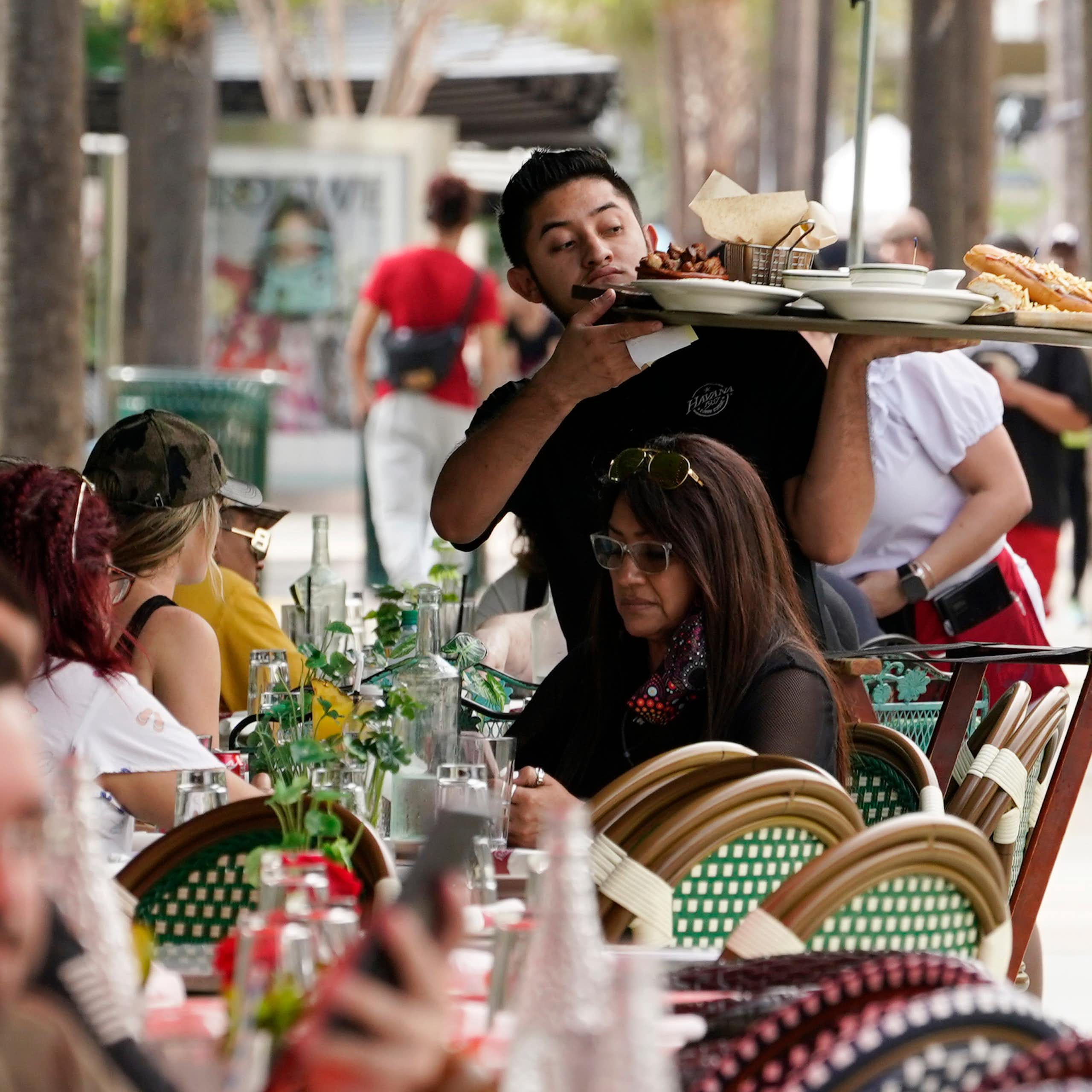 A server balances a large load of food on a tray above his customers' heads in an outdoor dining area.