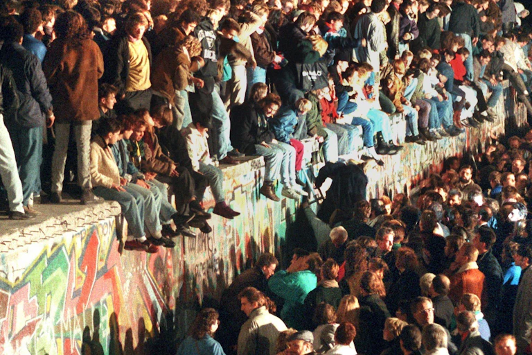 A historical photo of people standing and sitting on the Berlin wall as it is about to be torn down.
