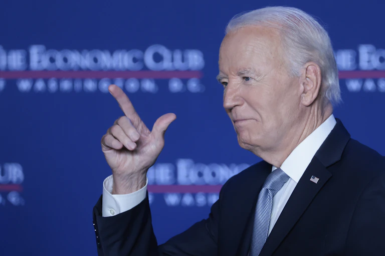 A man in a black suit points in the air while a backdrop says 'The Economic Club.'