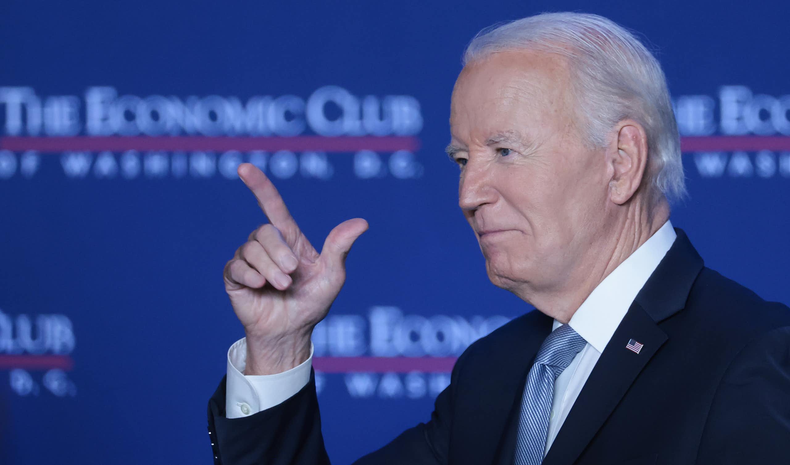 A man in a black suit points in the air while a backdrop says 'The Economic Club.'