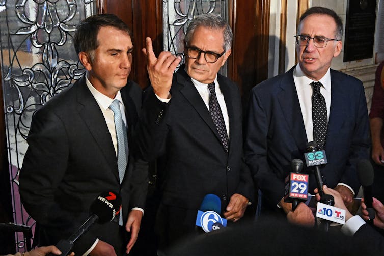 Three men in suits stand in front of a bunch of press microphones