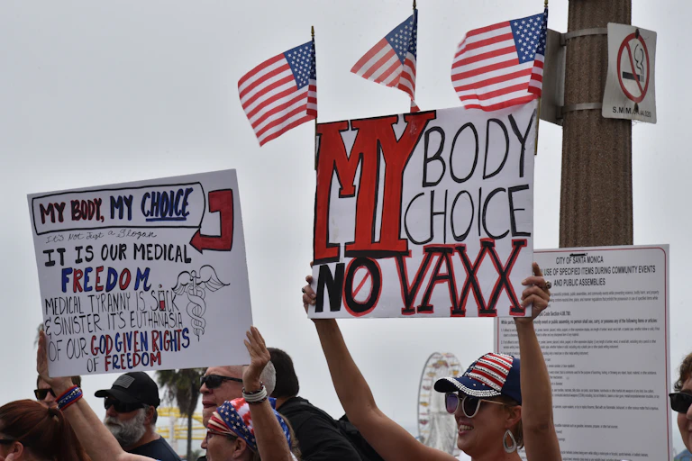 Protestors hold anti-vaccination signs against backdrop of US flags.