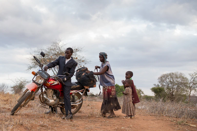 A man in a suit climbs onto a red and black motorcycle while a woman loads a bag onto the back and two small children look on