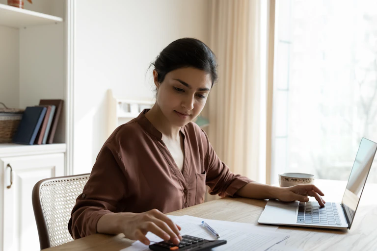 A young woman sitting at a table in her home, working on a computer and calculator