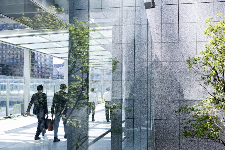 Two businesspeople are seen in silhouette walking through a glassy passage. They are framed by the reflections of leaves.