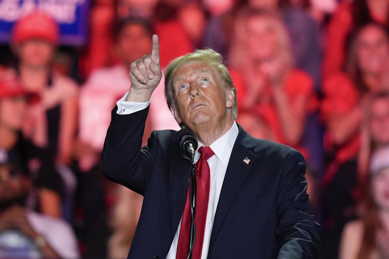 A man with unevenly applied face bronzer and greyish blond hair points up at the ceiling as supporters stand behind him.