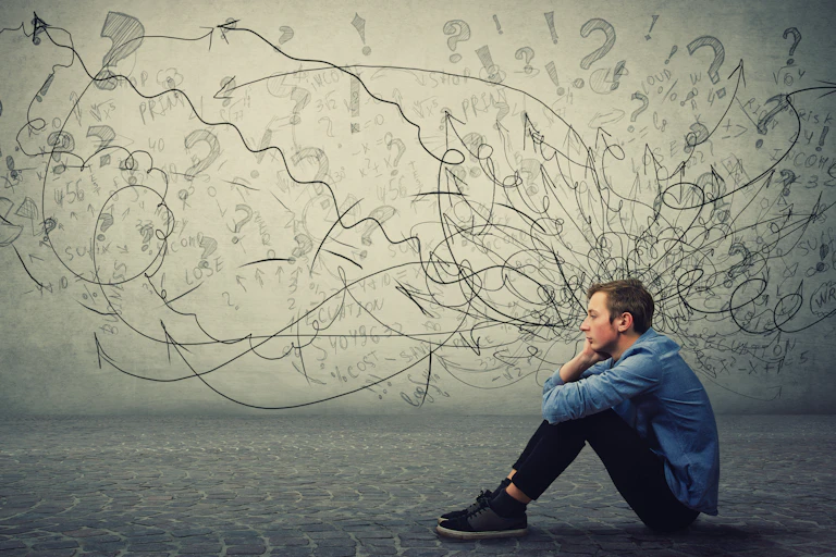 Young man sitting on the ground with lots of squiggles and question marks on a white wall behind him, suggesting this is happening in his head.