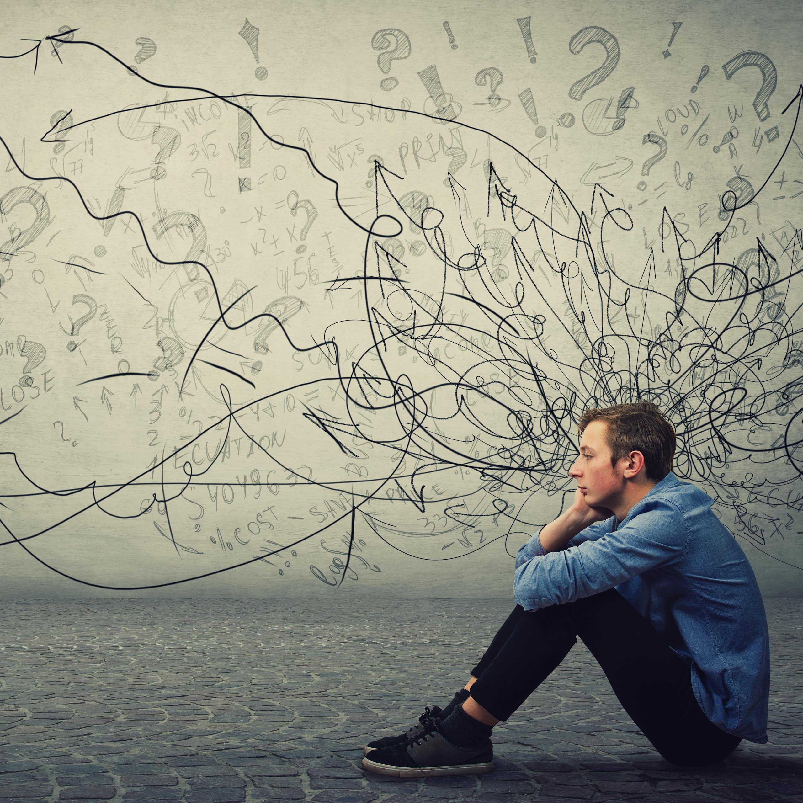 Young man sitting on the ground with lots of squiggles and question marks on a white wall behind him, suggesting this is happening in his head.