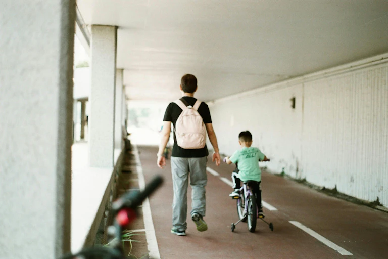 A man walks beside a young child riding a bike with training wheels.