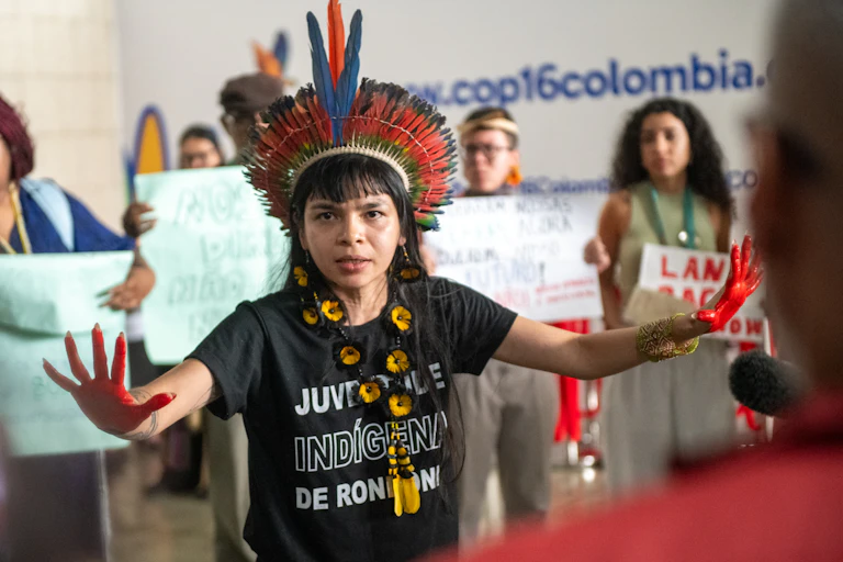 woman wearing black T shirt and colourful feathered traditional head dress, hands wide out, women holding signs in background