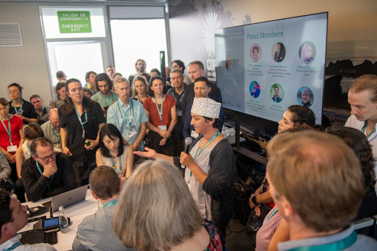 computer screen, people standing around listening to woman with microphone