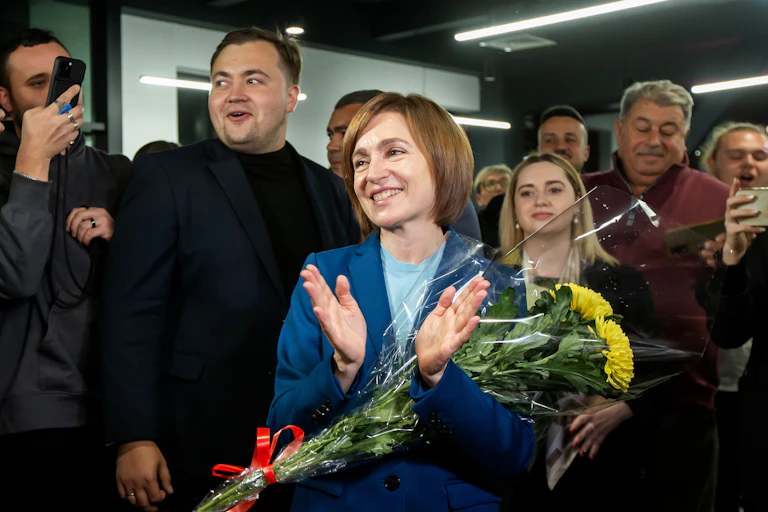 Newly reelected Moldovan president Maia Sandu, surrounded by supporters and carrying a sunfloewr, smiles and claps her hands.