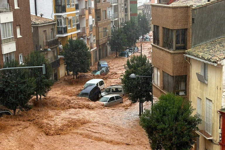 A photo taken from a high window of brown flood waters flowing through a town street, with cars being swept along by the current.