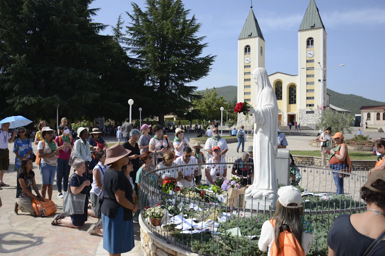 A white statue of the Virgin Mary stands against a backdrop of two tall towers. Many people stand around the statue, looking up and praying.