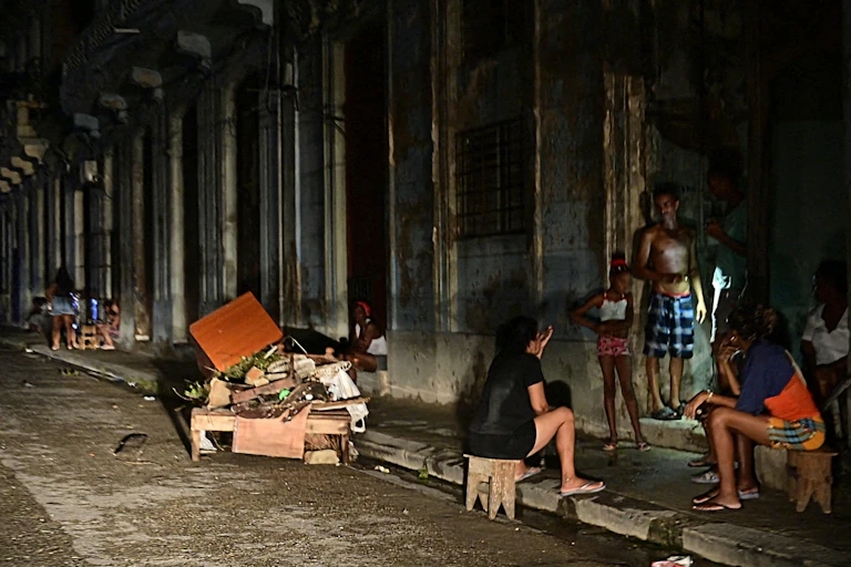 Men and women sit and stand on the side of a road, they are darkly lit.