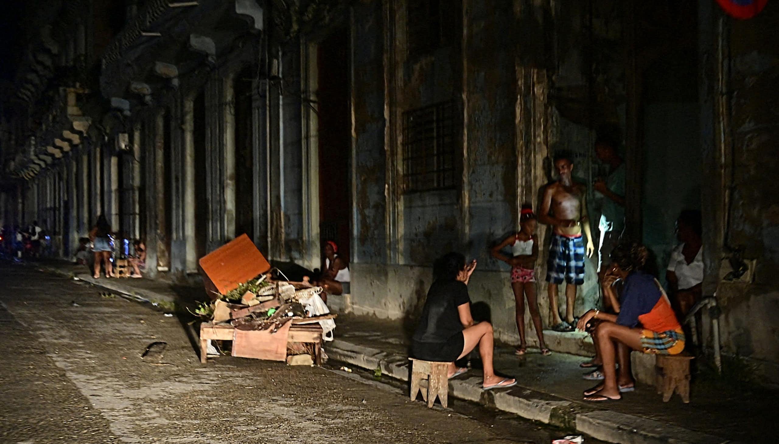 Men and women sit and stand on the side of a road, they are darkly lit.