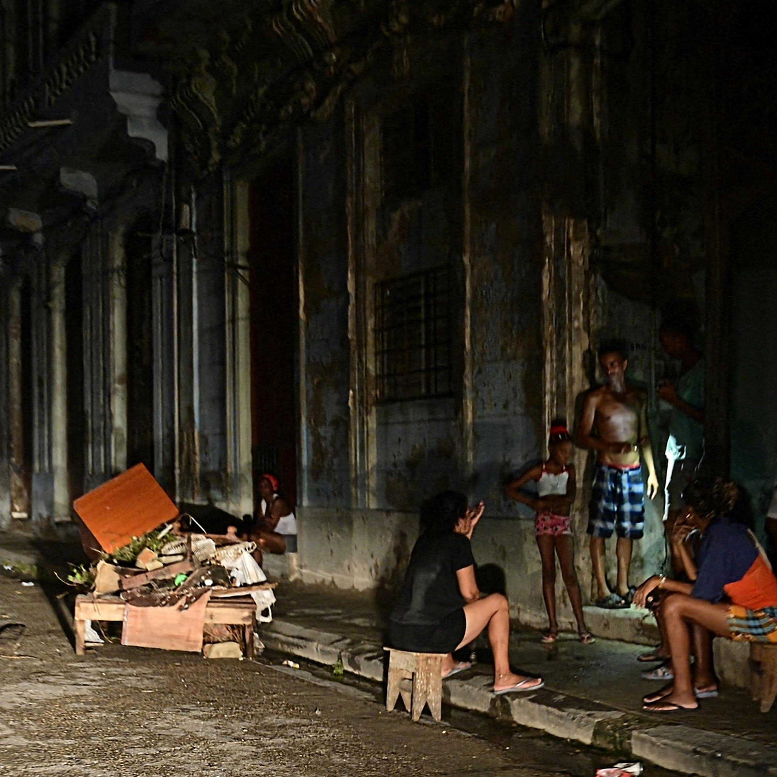 Men and women sit and stand on the side of a road, they are darkly lit.