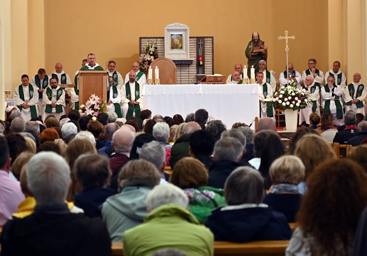Several men in priestly robes sit next to a table, covered with a white cloth, with lit candles on it and a crucifix to its side, while one person addresses the people seated in rows ahead.