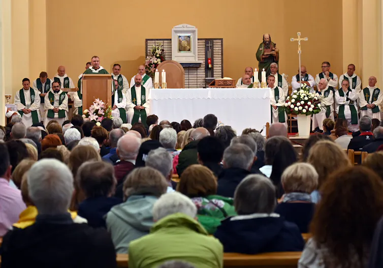 Several men in priestly robes sit next to a table, covered with a white cloth, with lit candles on it and a crucifix to its side, while one person addresses the people seated in rows ahead.
