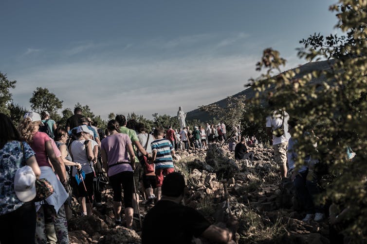 A long line of people walking on a steep, rocky path. In the distance is a white statue of the Virgin.