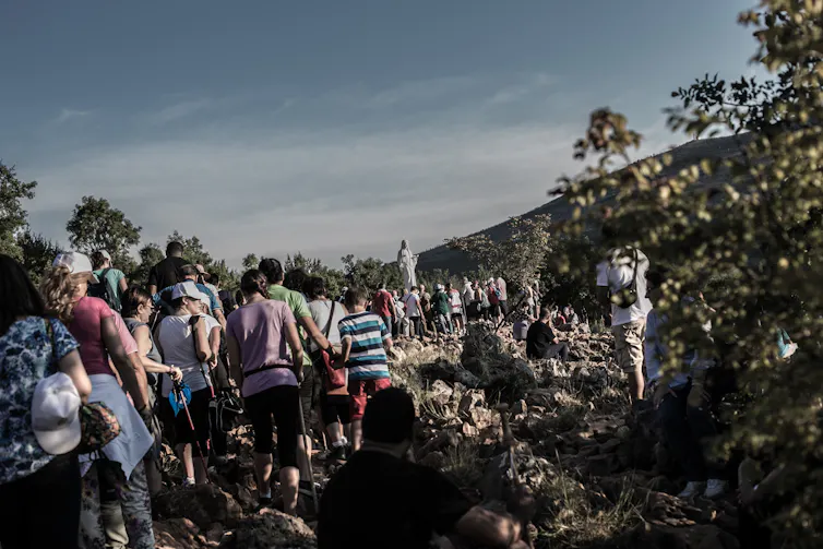 A long line of people walking on a steep, rocky path. In the distance is a white statue of the Virgin.