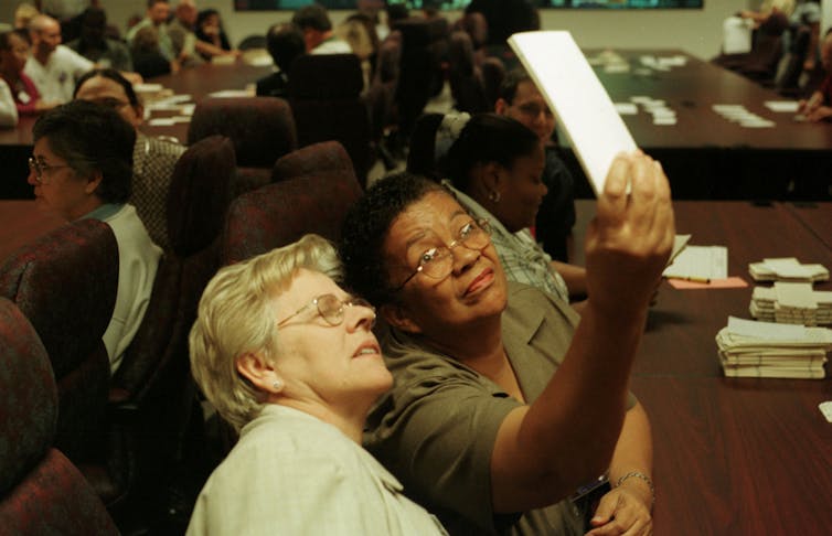 Two women wearing glasses sit at a table and hold up a white piece of paper and look at it.