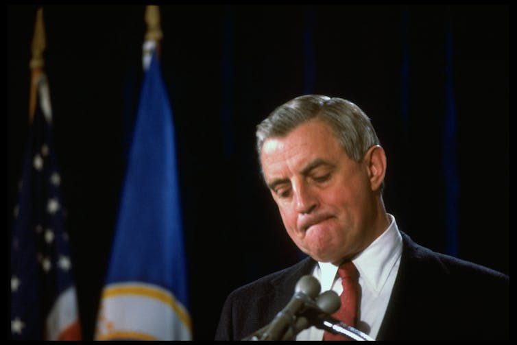 A white man wears a suit and looks down at a microphone, with part of the American flag visible behind him.