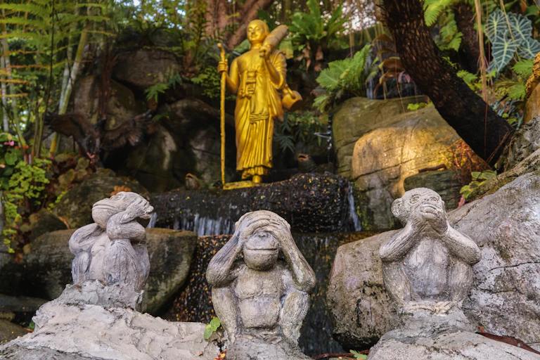 Gilded statue of Buddha as a walking monk with a Naga walking stick and umbrella and three wise seated monkeys' statues in foreground.