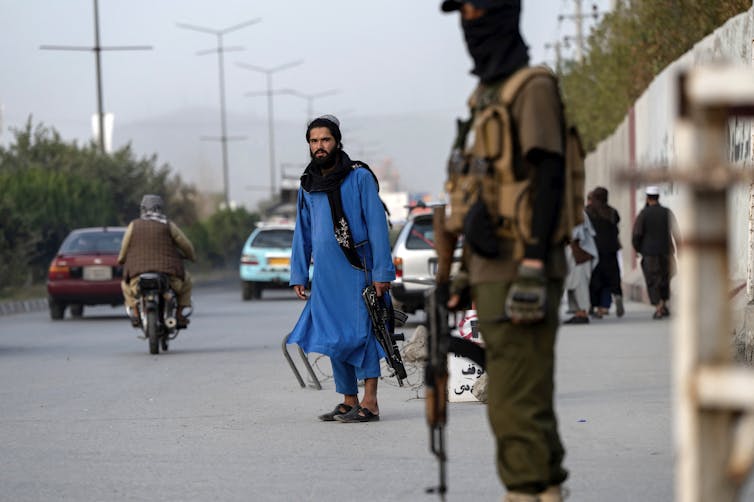 Armed soldiers stand near the entrance of a building as people and traffic can be seen in the background.