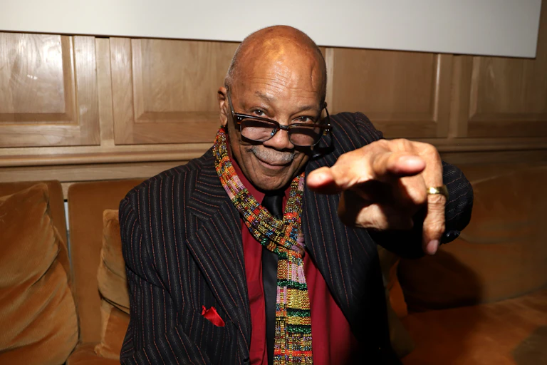 Bald black man wearing glasses points at a camera and grins.