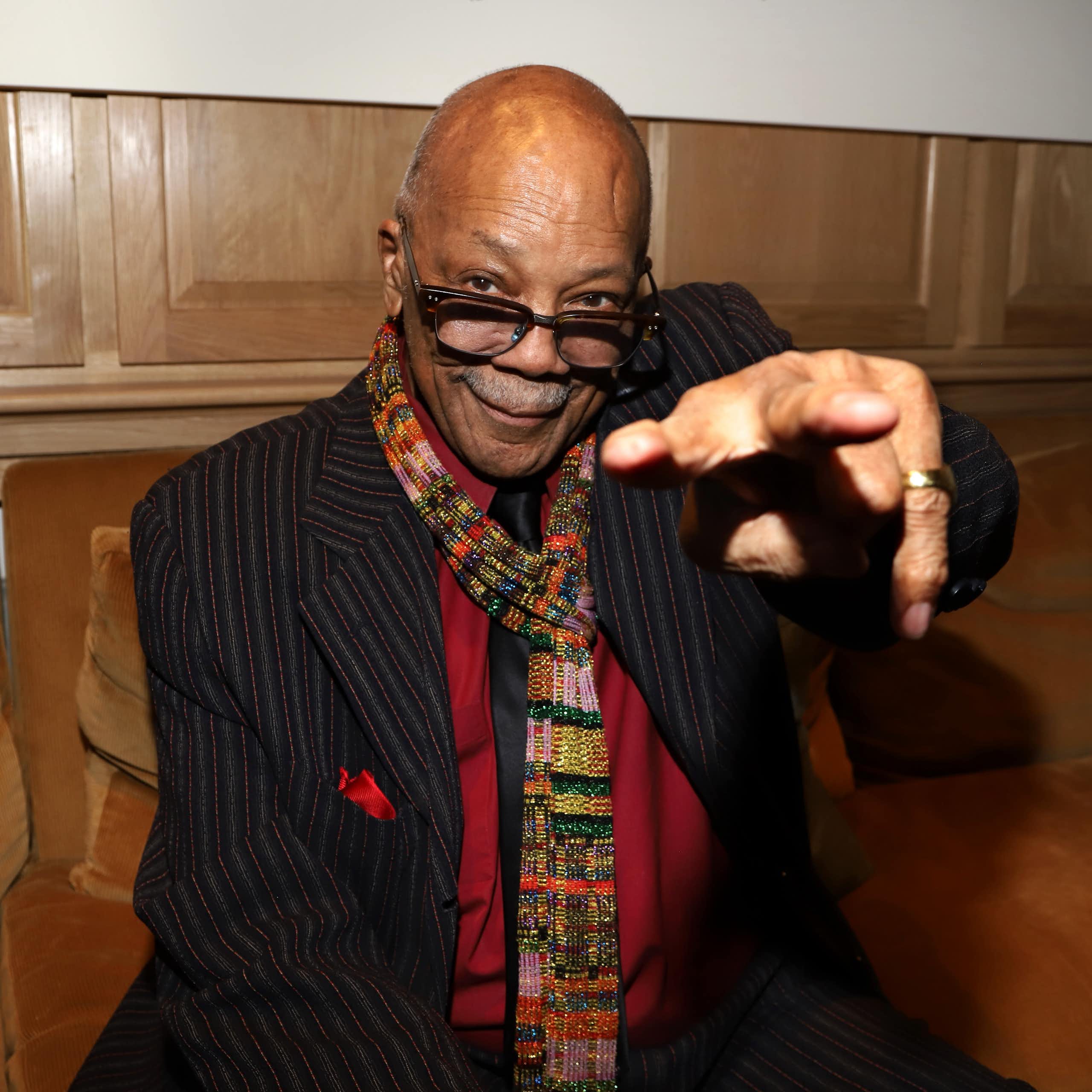 Bald black man wearing glasses points at a camera and grins.