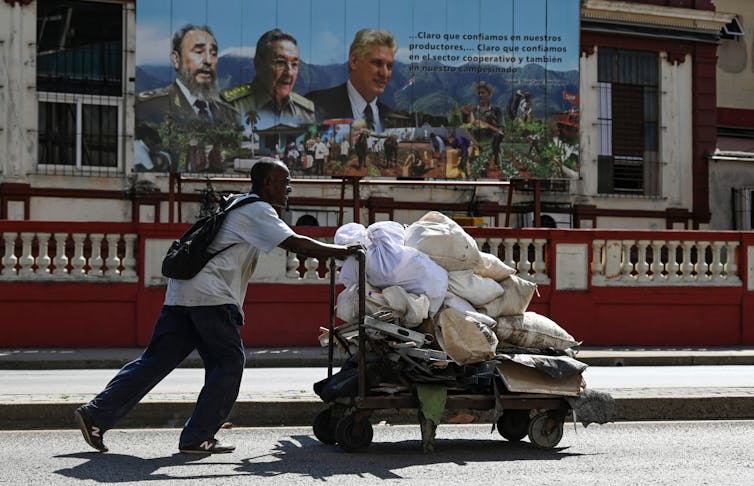 A man pushes a wheelbarrow of waste past a poster showing the image of three men.