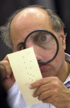 A man holds up a piece of paper with holes in it and inspects it with a magnifying glass.