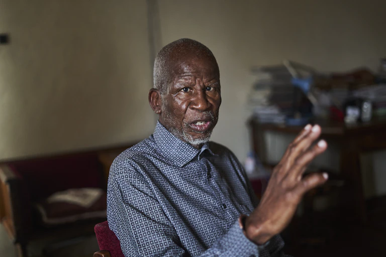An elder African man with a grey beard wears a blue collared shirt and gestures wirth one hand as he talks.