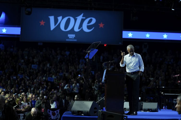 A Black man bows his head while on stage and points to a large banner that reads Vote.