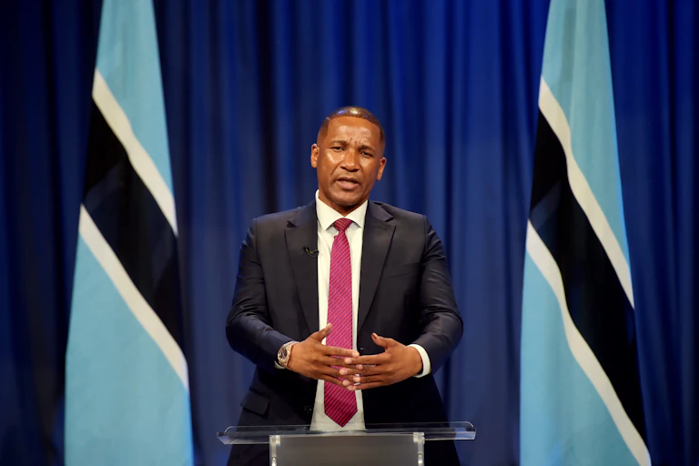 A man wearing a suit and tie speaks at a podium, in front of two flags of Botswana.