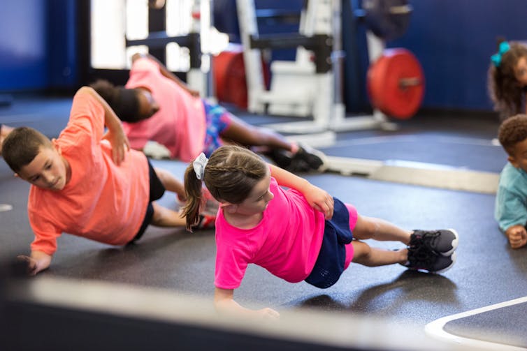 Group of kids performing side planks on mats in a gym.
