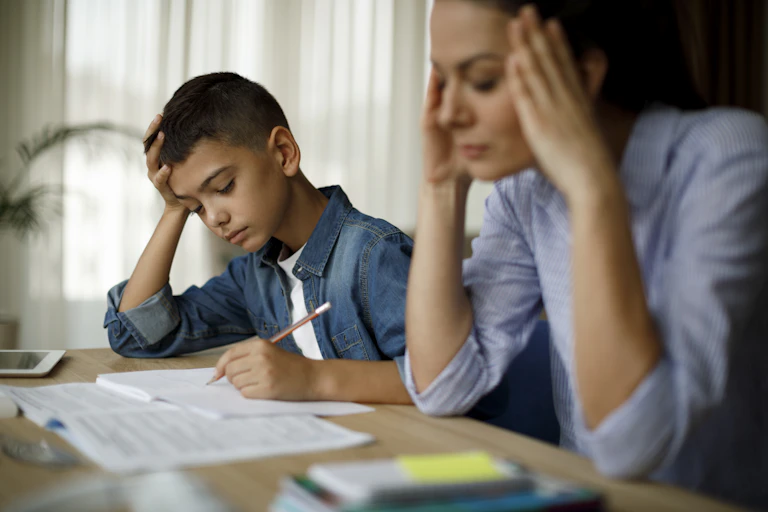 A frustrated mother places her hands on her head as to indicate a headache while her son struggles with his homework.