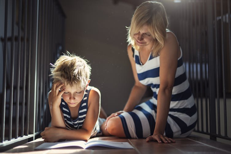 A mother helps her son do homework on the floor of a stairwell while sunlight shines in from an opening above.