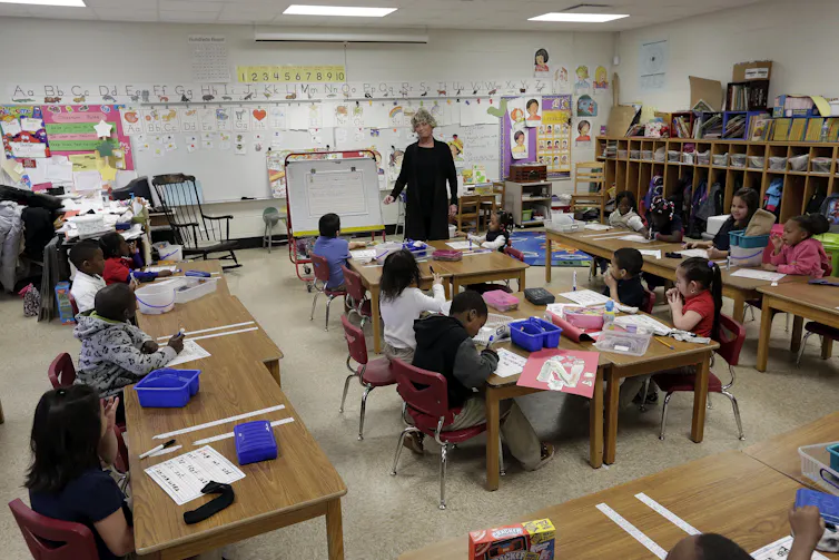 children in classroom sit at desks with teacher standing in background