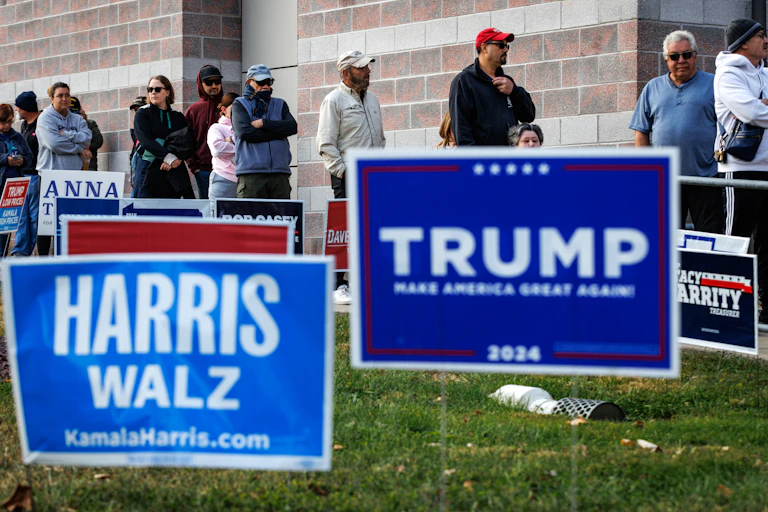 People stand in a line against a building. In front of them are large lawn signs, including two blue ones that say 'Harris Walz' and 'Trump.'