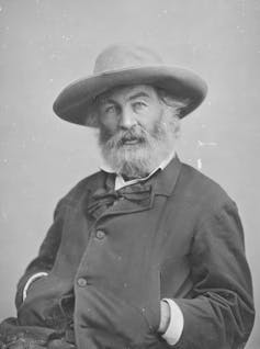 Black-and-white portrait of bearded man seated, wearing a floppy hat, with his hands in his jacket pockets.